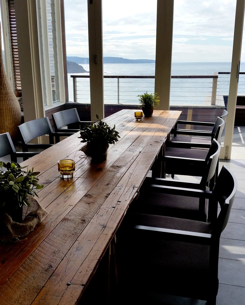 Vintage timber trestle table with black chairs on either side and a view of the ocean in the background