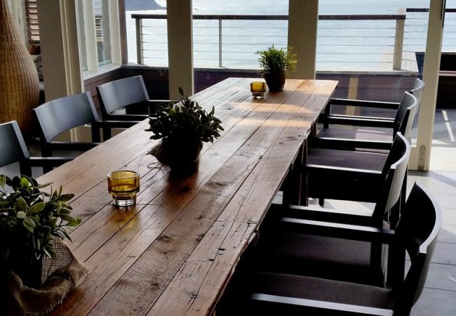 Vintage timber trestle table with black chairs on either side and a view of the ocean in the background